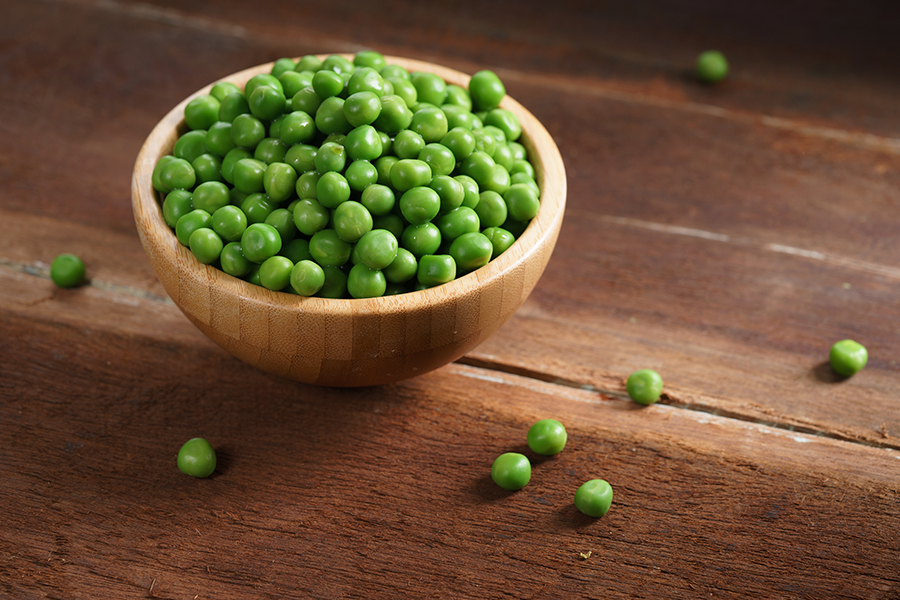 Um pequeno prato redondo de madeira cheio de ervilhas verdes brilhantes está sobre uma superfície de madeira, com várias ervilhas espalhadas ao redor.