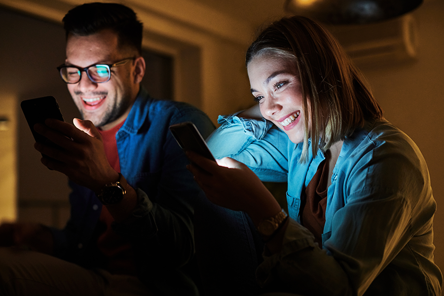 Um casal sorridente senta-se bem junto em uma sala escura, iluminada apenas pela luz das telas dos smartphones que estão olhando.