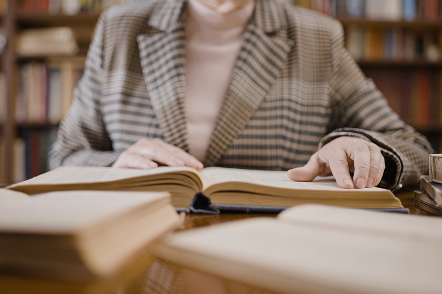 Una persona con una chaqueta a cuadros está sentada en un escritorio en una biblioteca, con las manos apoyadas en las páginas de un gran libro de tapa dura abierto.