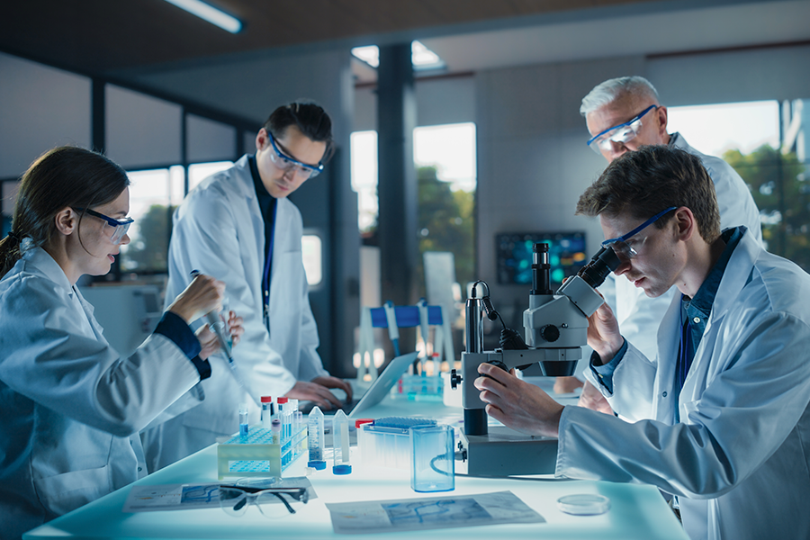 A diverse team of scientists in white lab coats and safety goggles work together in a modern laboratory using microscopes and pipettes.