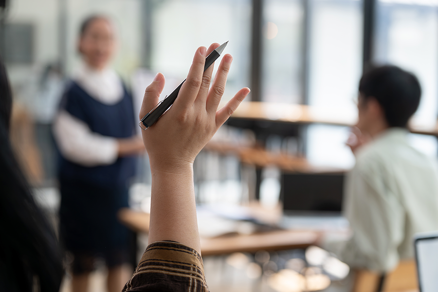 La mano de una persona sostiene un bolígrafo negro en alto para hacer una pregunta durante una presentación o una clase en el aula.