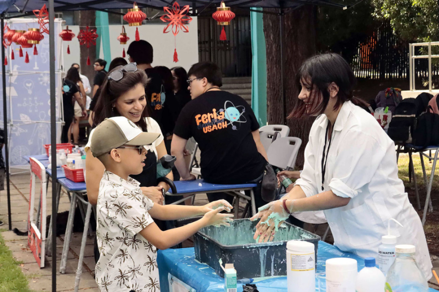 Científica Usach enseña a niño y su madre en un stand de la Feria de la Ciencia. La académica aparece con un delantal blanco y el niño cono un jockey café con blanco.
