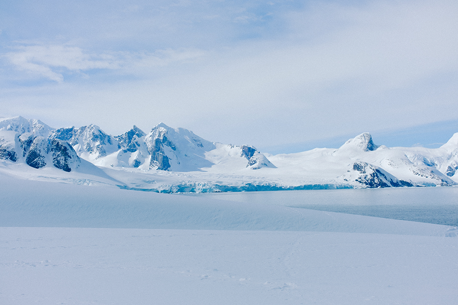 Un sereno paisaje de llanuras nevadas que conducen a montañas azules, escarpadas y heladas, bajo un cielo pálido.