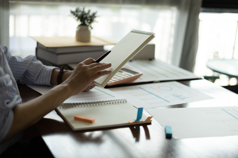 Una persona sentada ante un escritorio utilizando un bolígrafo y una calculadora sobre varios gráficos impresos y cuadernos abiertos.