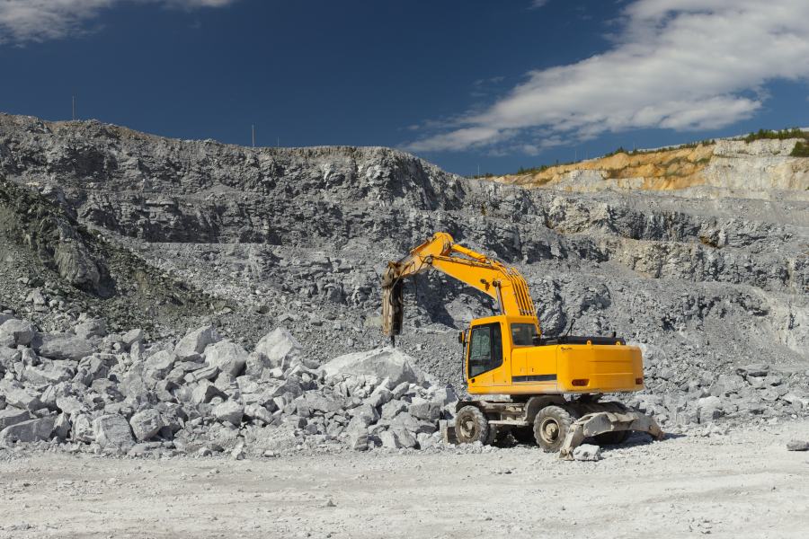 Yellow excavator with a hydraulic breaker working among piles of grey rocks in a wide quarry.
