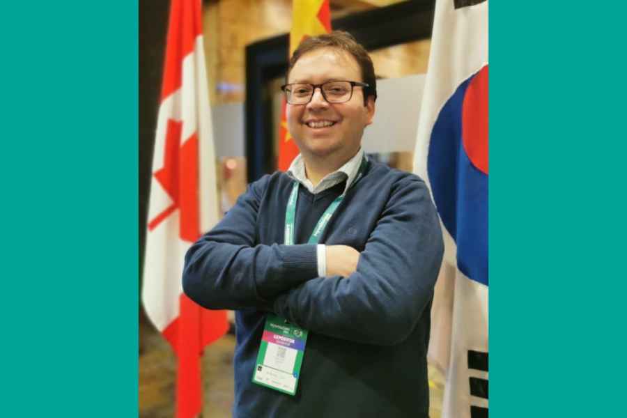 La imagen corresponde a una foto del académico posando de brazos cruzados, con tres banderas de fondo