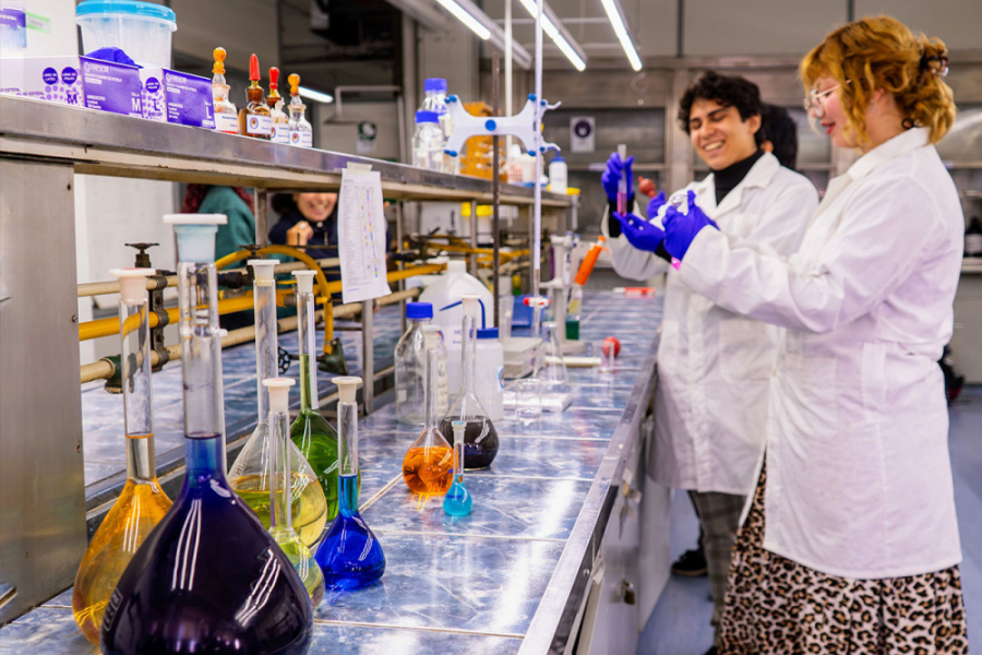 LA imagen muestra a dos estudiantes con cotona blanca y guantes azules trabajando en un laboratorio
