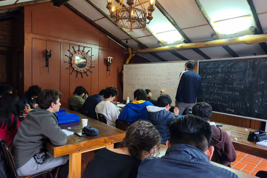 En la fotografía aparecen los estudiantes que participan del campeonato poniendo atención a un profesor dentro de una sala de clases. De fondo un pizarrón negro