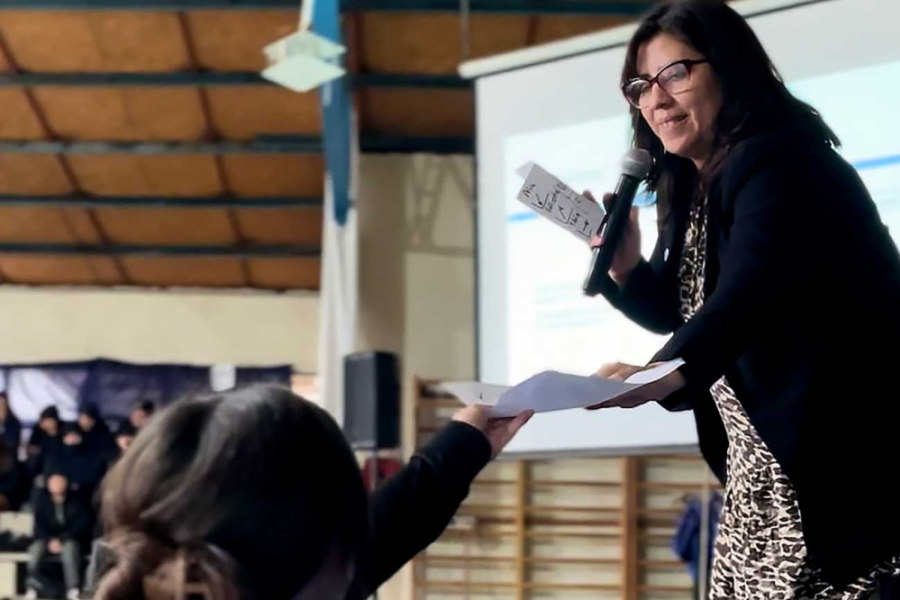 La imagen muestra a la mujer que dicta la charla, entregando un papel a una estudiante