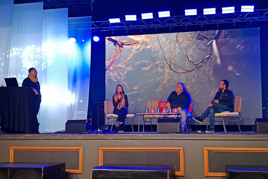 En la imagen se muestran a quienes expusieron en el Festival, sentados en el escenario. Donde hay una mujer con un micrófono en la mano y dos hombres escuchando