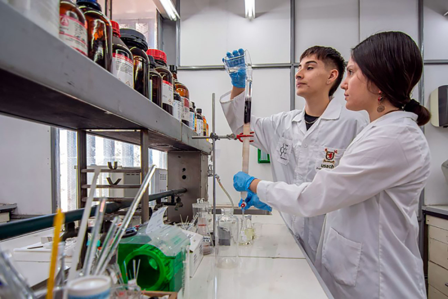 En la imagen aparecen dos estudiantes de la universidad trabajando en su laboratorio. Un hombre y una mujer que visten delantal blanco trabajan con pipetas y frascos.