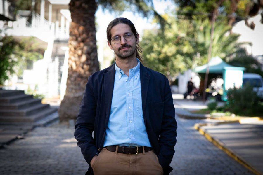 La imagen muestra al académico con las manos en los bolsillos, de pie frente a la cámara.