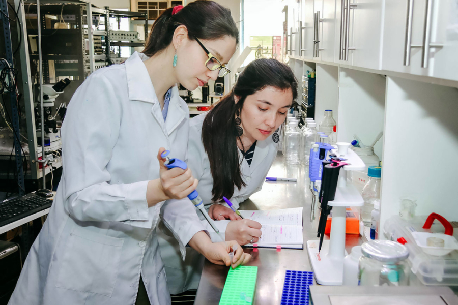 La imagen muestra a dos mujeres científicas con bata blanca en un laboratorio. Una manipula instrumentos mientras la otra registra lo observado