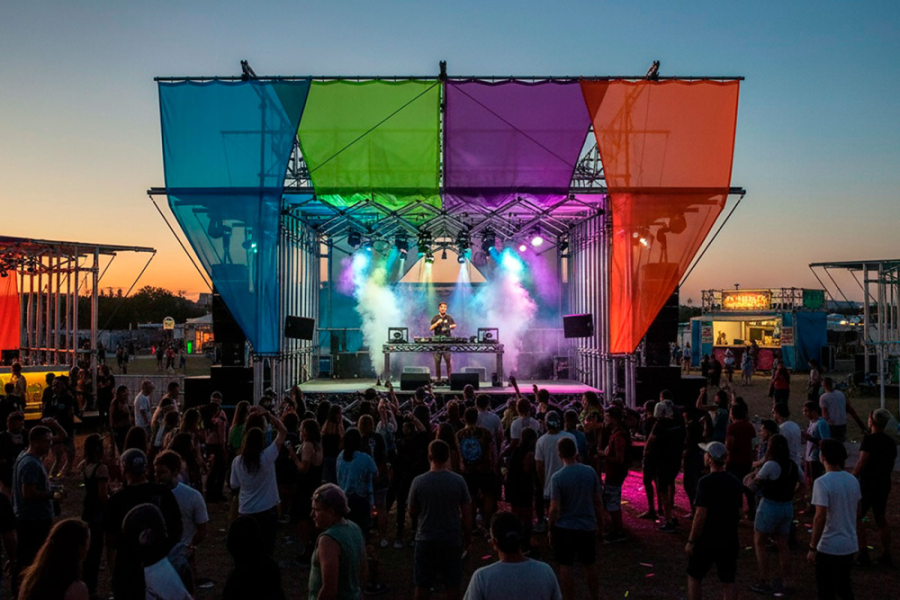 La foto muestra uno de los escenarios del evento. Con telas que adornan el espacio de colores celeste, verde, morado y naranjo y un hombre ubicado al centro del escenario con una mesa de sonido