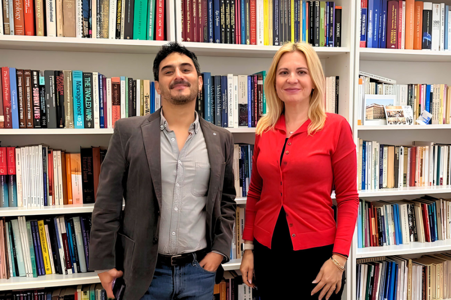 En la fotografía aparece el Dr. David Jofré Leiva, académico de la Escuela de Periodismo que realizó una movilidad en Rumania. En la fotografía aparece delante una gran biblioteca, junto a una colega de la Universidad de Transilvania de Braşov. ella vestida de rojo y él con chaqueta y camisa gris.