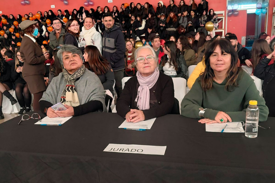 En la imagen aparecen el primer plano las tres jurados del evento. Están sentadas y sus manos cruzadas sobre una mesa. Detrás de ellas el público general