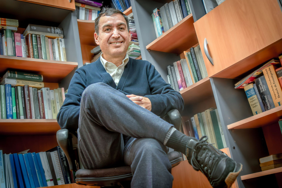 En la fotografía aparece el Dr.Carlos Lizama, investigador del Departamento de Matemática y Ciencia de la Computación de la Usach, lidera un Fondecyt Regular 2026 . está sentado en u na silla con las piernas cruzadas. De fondo una gran biblioteca con libros.