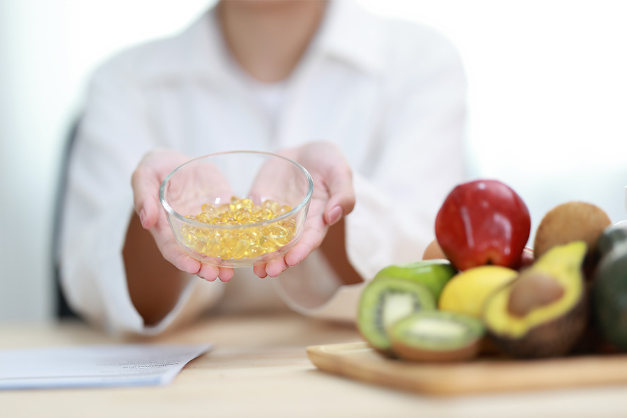 Persona sosteniendo un recipiente con cápsulas de gel junto a frutas y verduras frescas.