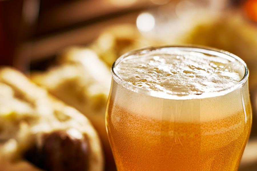 A glass of lager with lots of foam in the foreground and food blurred in the background.