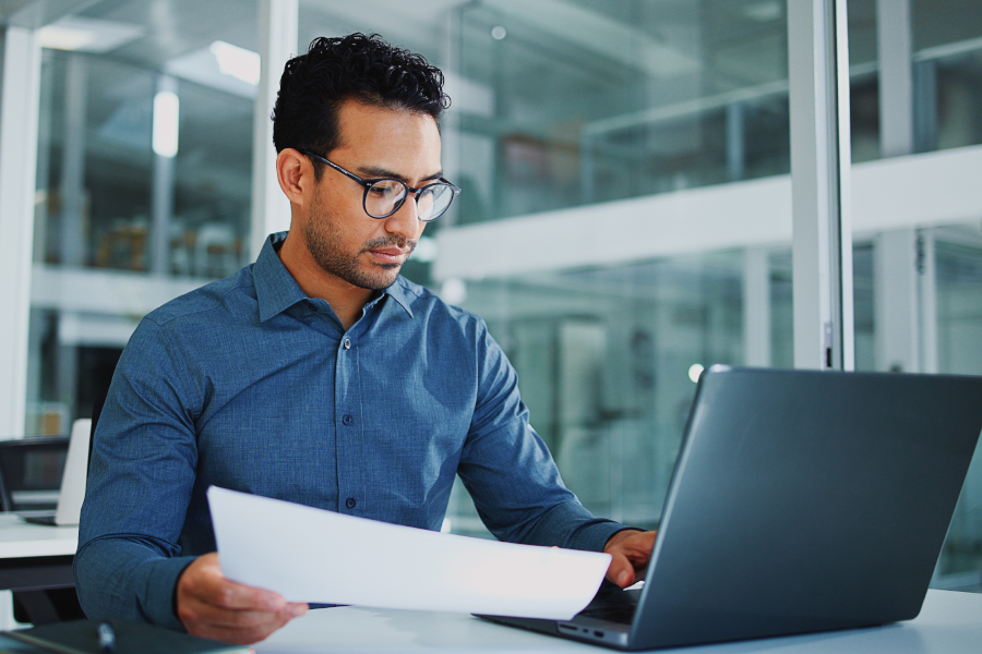 Man with glasses reviewing a document while working on laptop.
