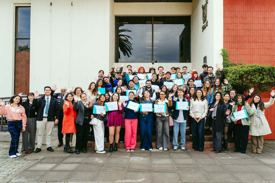 Grupo de estudiantes y autoridades posa frente a un edificio del campus. Algunas personas sostienen certificados y varias levantan la mano en señal de saludo mientras miran a la cámara