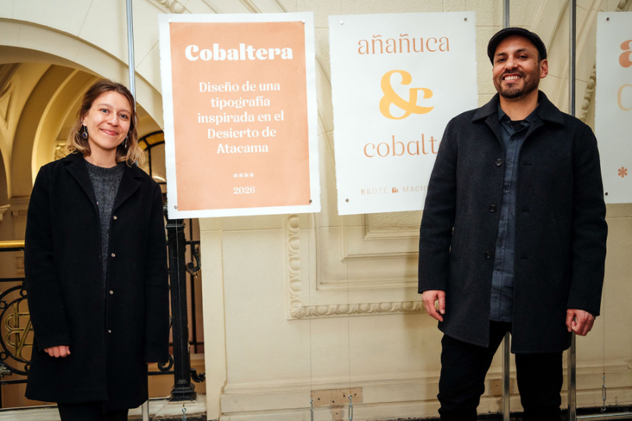 La imagen muestra al docente junto a la diseñadora, posando frente a los posters de la muestra