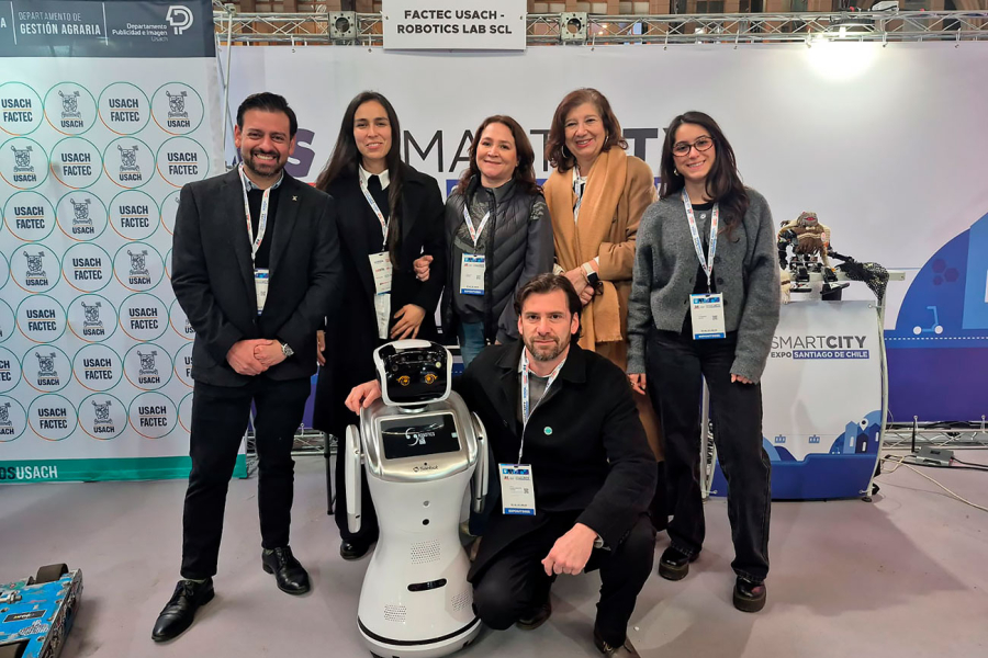 Seis personas posan junto a un robot de servicio blanco y plateado en el stand de Factec Usach y Robotics Lab SCL durante la Expo Smart City. Al fondo se observan paneles con los logos institucionales y la gráfica del evento. El grupo sonríe a la cámara 