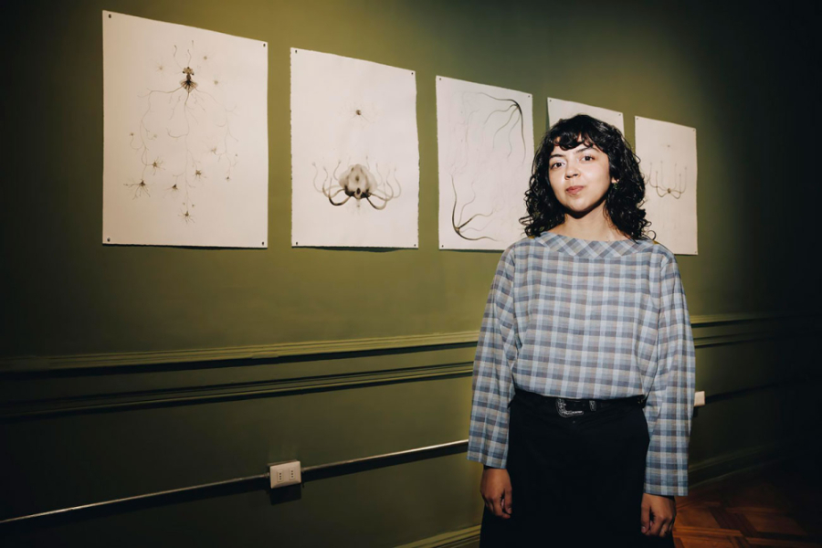 La foto muestra a la artista visual posando frente a una de sus exposiciones. De pie con una blusa a cuadros y pantalones negros, de fondo la pared verde y cinco de sus obras