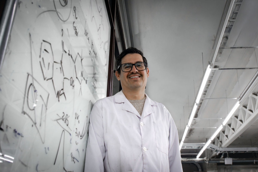 La imagen muestra al académico, de bata blanca, sonriente, apoyado en una pizarra de su laboratorio