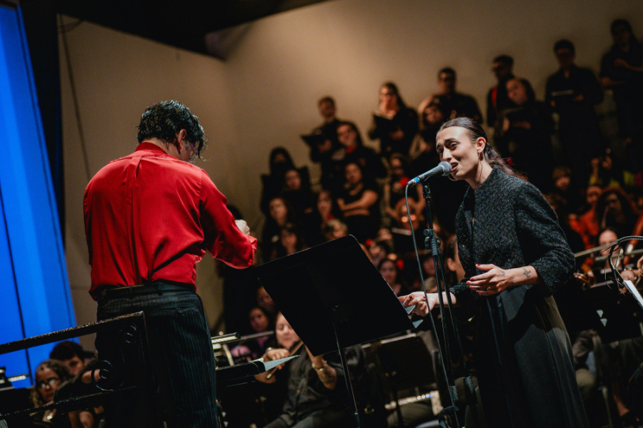 En la imagen aparece el director de la Orquesta Usach dirigiendo al elenco. Aparece con una camisa roja.  A su lado una intérprete que aparece vestida de negro cantando frente al micrófono. De fondo los músicos de la Orquesta Usach