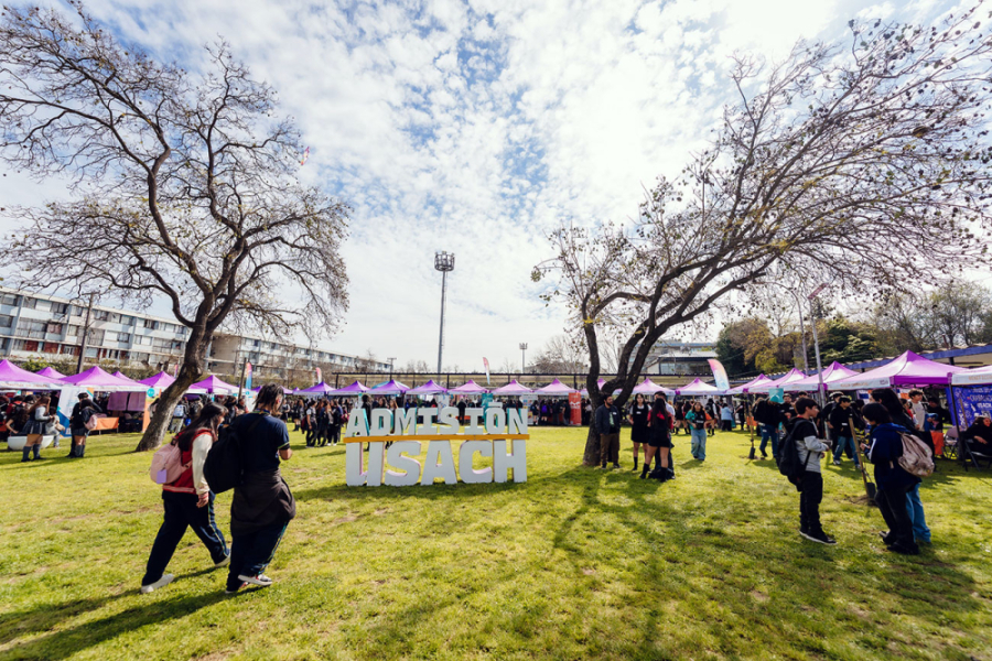 En la imagen aparecen estudiantes de los diversos colegios que visitaron la muestra que se instaló en los pastos de ciencia. En medio de la imagen aparecen las letras institucionales "Admisión Usach"