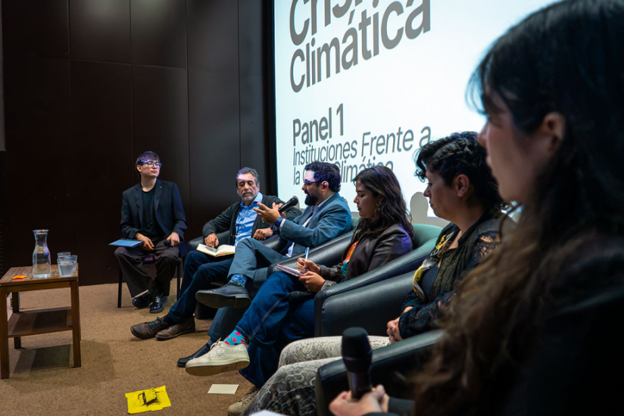 En la imagen aparecen los participantes del panel de crisis clímatica. Son tres hombres y tres mujeres. Están sentados y detrás de ellos una enorme pantalla led con el nombre del conversatorio.